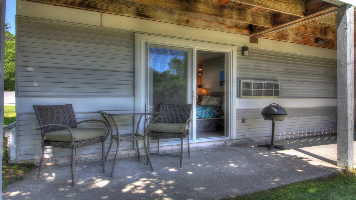 A small and inviting outdoor patio space with chairs, a table, a grill, and a sliding glass door leading directly into the suite. The setting is perfect for quiet mornings or social evenings, blending comfort and connection with nature.