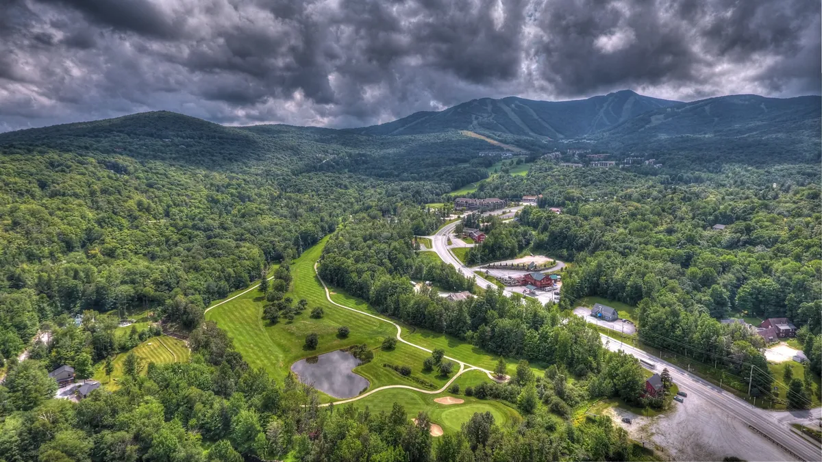 A panoramic view of a lush, green golf course winds alongside a gentle river, framed by distant mountain peaks. The landscape is fresh and inviting, suggesting opportunities for both relaxation and active outdoor pursuits like golfing, hiking, and nature walks.