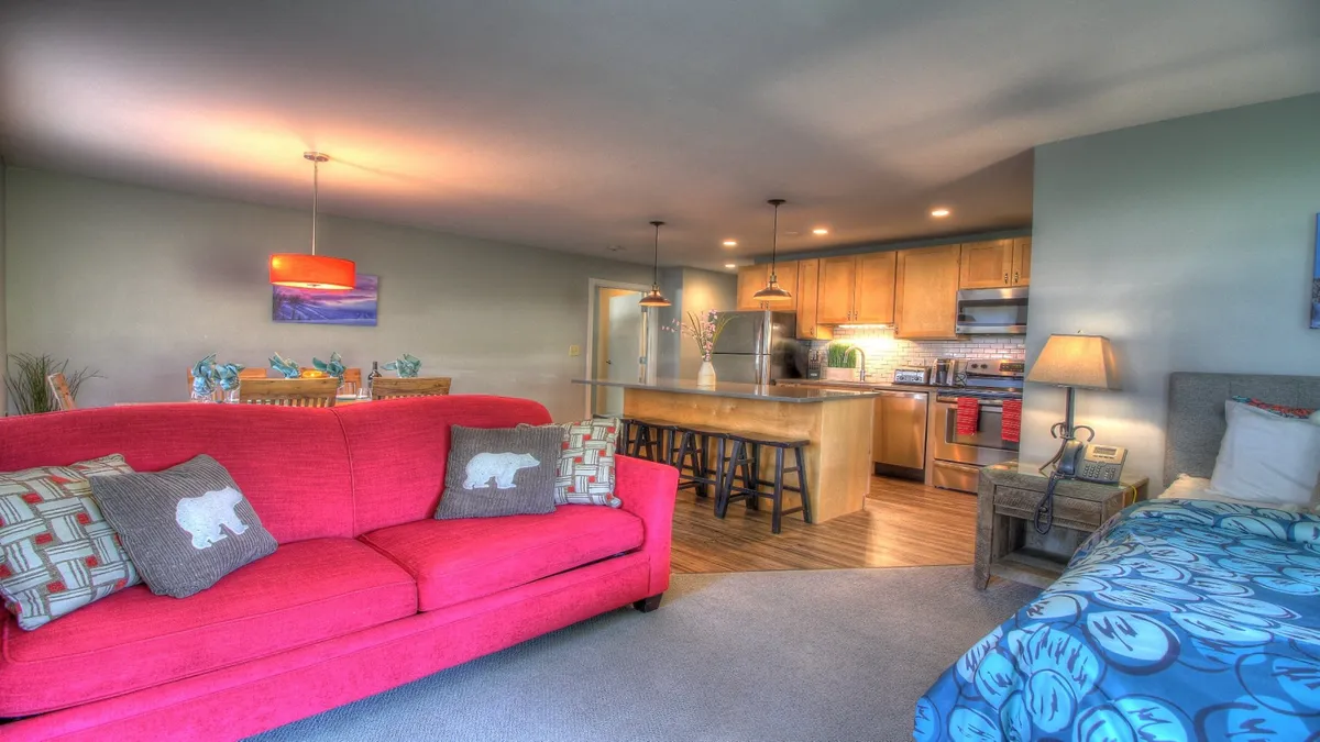 This image features a seamless open-concept layout encompassing a red sofa, a spacious kitchen island, and a bed with patterned bedding. The kitchen area includes stainless steel appliances and the island doubles as a gathering spot, visually connecting the living, dining, and kitchen spaces. The setup suggests flexibility for both cooking and relaxing, mirroring the multi-use nature of the suite.