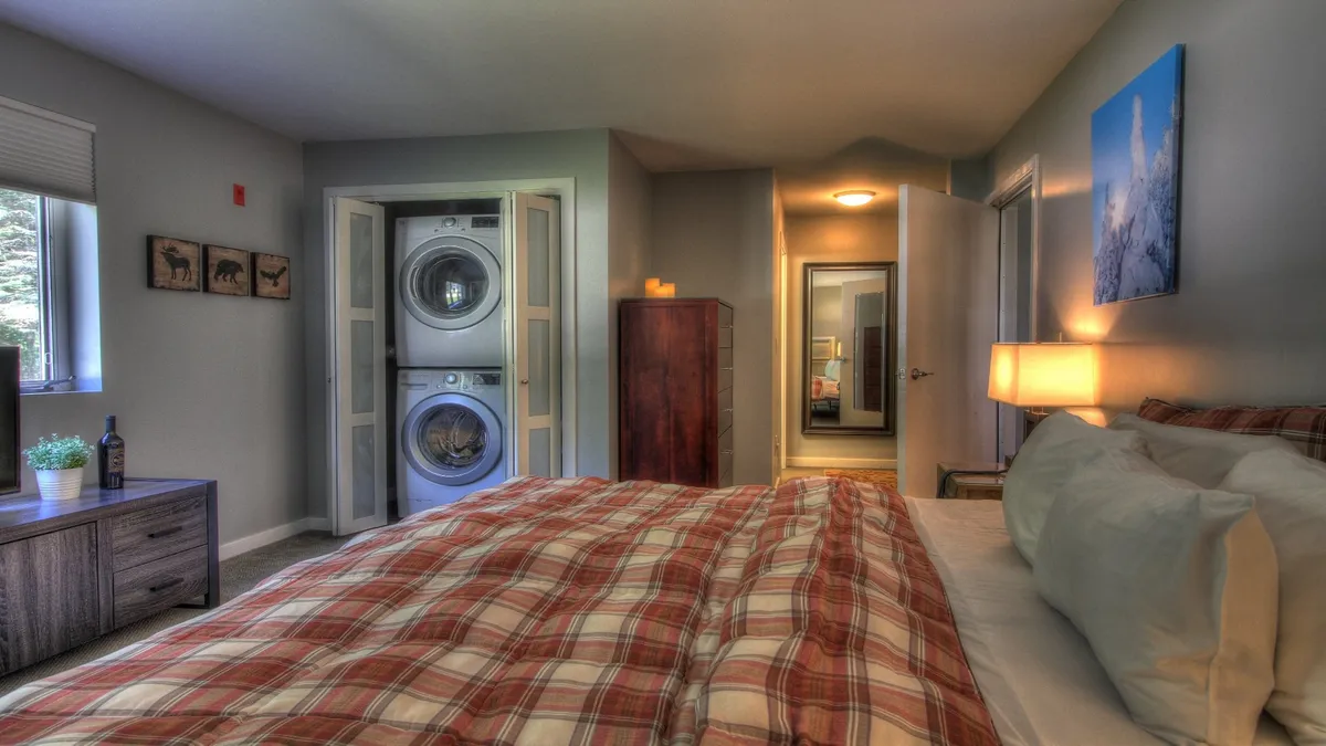 A cozy master bedroom with plaid bedding, a wooden nightstand, framed artwork, and an in-suite washer and dryer discreetly positioned to the side. The room's inviting ambiance highlights practical features for longer stays.
