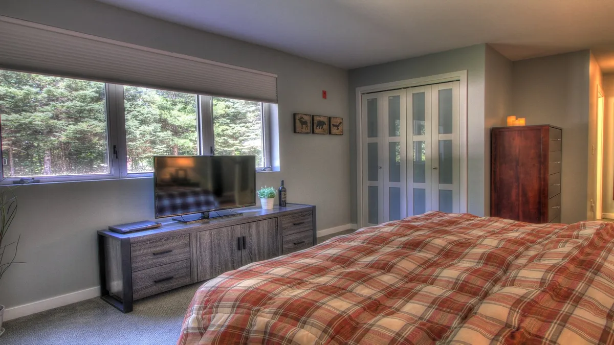 A calming bedroom interior featuring a bed, large windows allowing natural light to filter in, a dresser, and a flat-screen TV positioned for easy viewing from the bed.