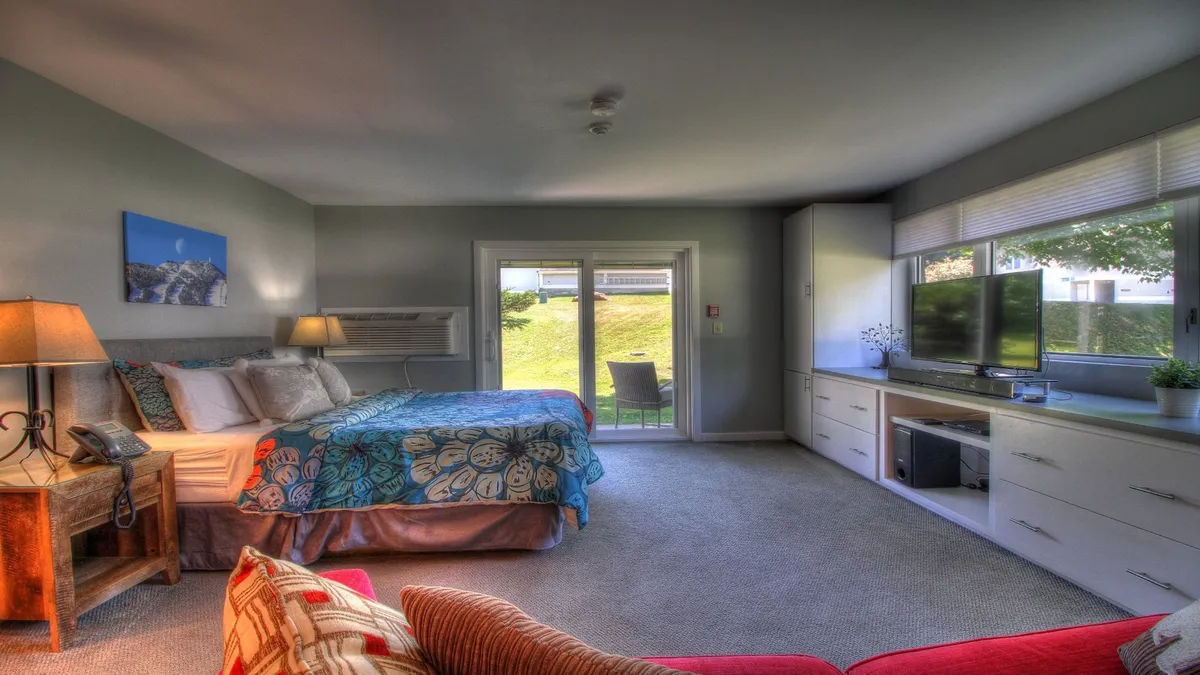 A cozy suite with a bed covered in colorful bedding, a TV set on a white cabinet, a red accent chair, and sliding glass doors that open to an outdoor space. The space is bright with natural light, suggesting an easy transition between relaxing indoors and enjoying the outdoors.