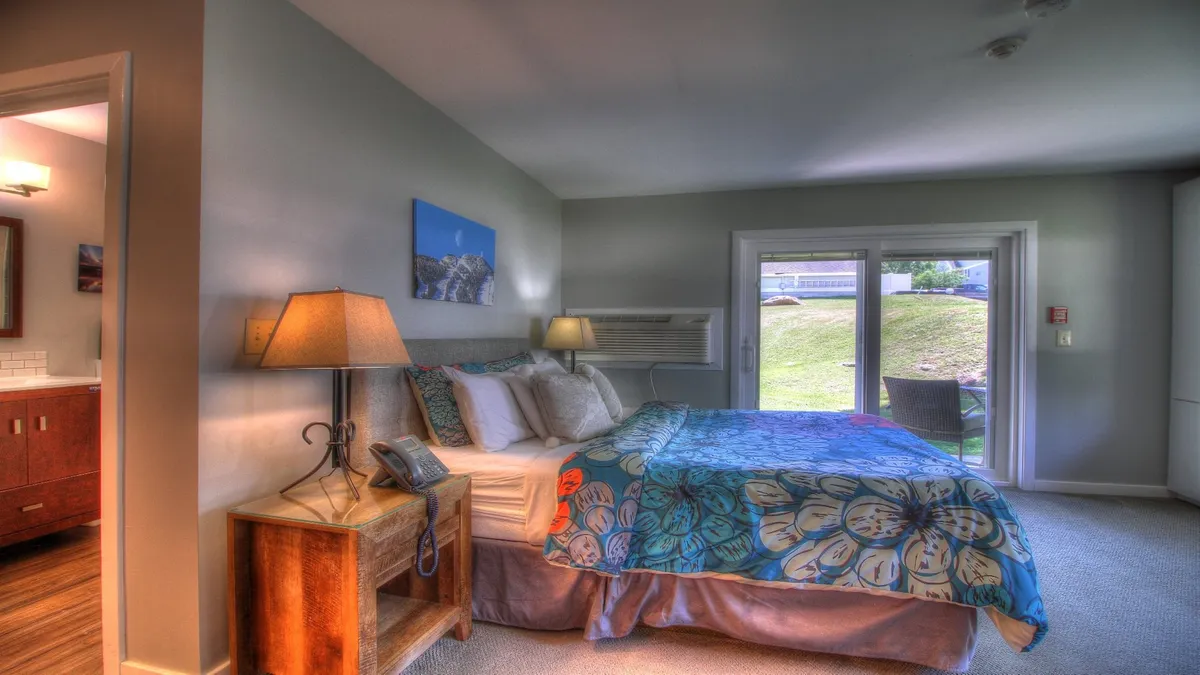 This image shows a cozy bedroom with colorful bedding, a lamp on the bedside table, and a view through sliding glass doors that likely lead to a private patio. Natural light streams in, and the scene suggests easy indoor-outdoor living for guests who want to sip coffee by the patio or enjoy the fresh air.